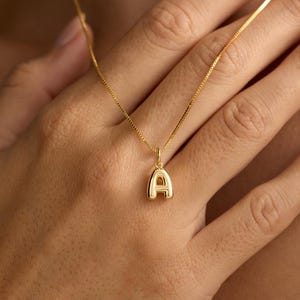 May include: A gold-toned necklace featuring a small, puffed letter A pendant suspended from a delicate box chain. The pendant has a smooth, rounded surface and a polished finish, reflecting light across its curved, three-dimensional shape.