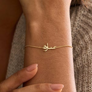 May include: A delicate gold chain bracelet featuring a central nameplate with Arabic script. The thin, polished metal chain connects to the custom lettering, creating a refined piece of jewellery that rests horizontally across the wrist area.