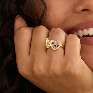 May include: A gold-toned heart-shaped locket ring featuring a hinged design that opens to reveal a small photograph of people inside. The ring has a polished metallic finish and is worn on a finger, showcasing the functional locket mechanism.