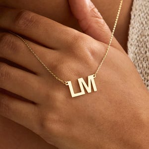 May include: A gold-coloured necklace featuring a pendant with the capital letters L and M in a block font. The pendant is attached to a delicate, thin gold-coloured chain that rests against the skin. The letters are connected in a single piece.