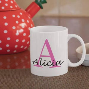 May include: White ceramic mug with a pink letter "A" and the name "Alicia" in a script font. A red teapot with white polka dots is in the background. The mug is on a brown surface.