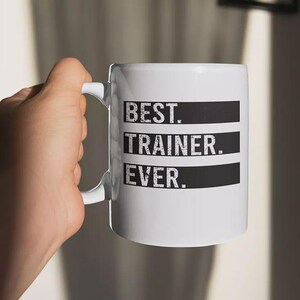 May include: White ceramic mug with the text "BEST. TRAINER. EVER." in black, block letters. The text is arranged in three lines, each on a black rectangular background. The mug has a white handle.