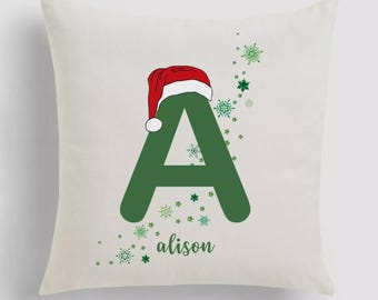 Könnte beinhalten: Cremefarbenes Zierkissen mit einem großen grünen Buchstaben "A", bedeckt mit einem rot-weißen Weihnachtsmannhut. Der Name "alison" ist in grüner Schrift darunter gedruckt, mit grünen Schneeflocken-Akzenten.