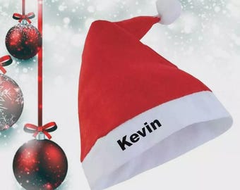 May include: A red Santa hat with a white brim and a white pom-pom. The name "Kevin" is printed in black on the white brim. Christmas ornaments hang in the background.