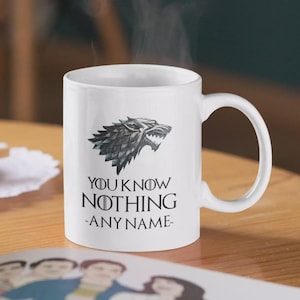 Pode incluir: Caneca de cerâmica branca com um emblema de lobo gigante e o texto "YOU KNOW NOTHING -ANY NAME-". A caneca tem uma alça curva e é destinada a bebidas quentes. O vapor sobe da caneca.