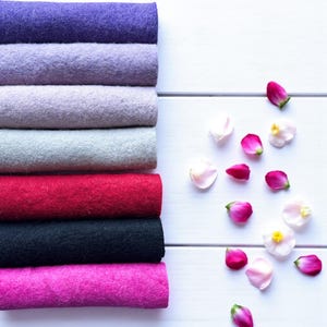 May include: A stack of colorful felt fabric rolls in shades of purple, gray, red, black, and pink. The felt is arranged next to scattered pink and white flower petals on a white wooden surface.