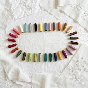 May include: A collection of colorful thread spools arranged in an oval shape on a white, textured surface. The spools display a variety of colors, including pink, blue, green, and brown. The background features white fabric.