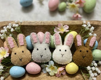 May include: Four felt bunny heads in shades of brown, gray, and white, each with pink-lined ears, are arranged in a wooden tray. Pastel-colored Easter eggs and small flowers surround the bunnies, creating a festive spring display.