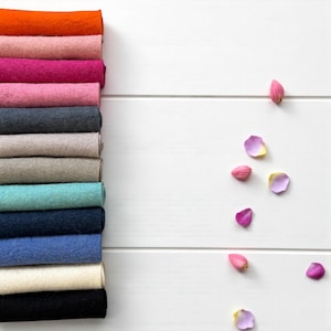 May include: A collection of felt fabric rolls in various colours, including orange, pink, and blue. The rolls are stacked on the left against a white wooden backdrop, with scattered pink and purple flower petals.