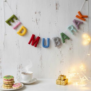 May include: A colorful felt banner spelling "EID MUBARAK" hangs against a white wooden background. The letters are in various colors, strung together with white string. A cup of tea, cookies, and small gold gift boxes are on a table below.