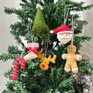 Könnte beinhalten: Handgefertigte Weihnachtsschmuckstücke hängen an einem grünen Weihnachtsbaum. Die Ornamente umfassen einen grünen Filz-Weihnachtsbaum, einen Weihnachtsmannkopf, einen Lebkuchenmann, ein Rentier und eine Zuckerstange.