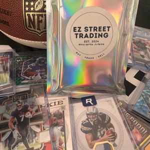 May include: An assortment of football trading cards, including rookie cards, and a holographic bag with the text "EZ STREET TRADING". A brown football with the NFL logo is visible in the background.