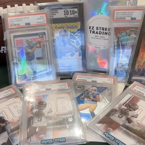 May include: A collection of graded football trading cards in protective cases, featuring images of players in action. A brown NFL football is visible in the background. The cards are from various brands and include player names and grading information.