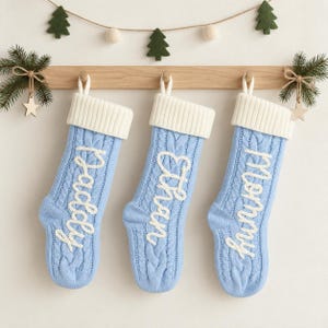 Puede incluir: Tres calcetines navideños de punto trenzado en color azul claro colgados de una barra de madera. Cada calcetín tiene un puño acanalado blanco y texto bordado en blanco con los nombres Daddy, Ethan y Mommy en vertical.