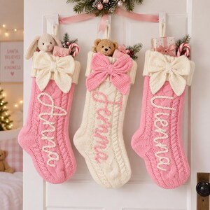 May include: Three cable-knit Christmas stockings hanging on a white door. The left and right stockings are pink with white names Anna and Vienna, while the centre is cream with the name Jemma in pink. Each features a large bow and small toys.