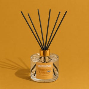 May include: A clear glass reed diffuser bottle with a gold-coloured collar and six black reeds. The label features the text PEACH THREADS and SANDALWOOD & VANILLA in white and gold lettering against a light orange background on the glass.