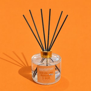 May include: A clear glass reed diffuser bottle with a gold-coloured collar and six black reeds. The label features the text "PEACH" repeatedly at the top, followed by "SICILIAN LIME, MANDARIN & BASIL" and "PETAL SWEET & CRISP" in orange and white.