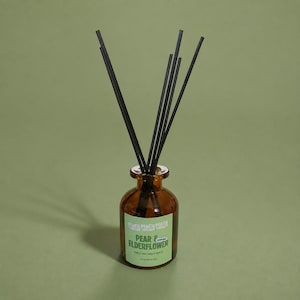 May include: A reed diffuser featuring a small amber glass bottle with a light green label. The label displays the text "PEACH PEACH PEACH" at the top and "PEAR & ELDERFLOWER" in large green letters, with "PEAR, ELDERFLOWER & VANILLA" below. Six black reeds extend upwards.