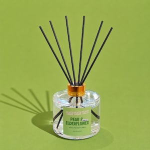May include: A clear glass reed diffuser with a gold-coloured collar and six black reeds. The label on the front features the text PEACH THREADS, PEAR & ELDERFLOWER, and PEAR, ELDERFLOWER & VANILLA, with a small label reading PREMIUM SCENT.
