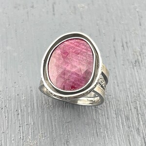 May include: A silver ring featuring a large, oval-shaped, faceted pink gemstone set in a smooth metal bezel. The band of the ring is constructed from multiple thin, layered silver wires that create a textured, openwork appearance along the sides.