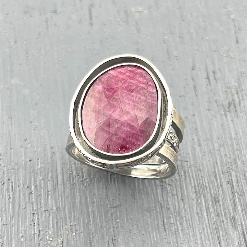 May include: A silver ring featuring a large, oval-shaped, faceted pink gemstone set in a smooth metal bezel. The band of the ring is constructed from multiple thin, layered silver wires that create a textured, openwork appearance along the sides.