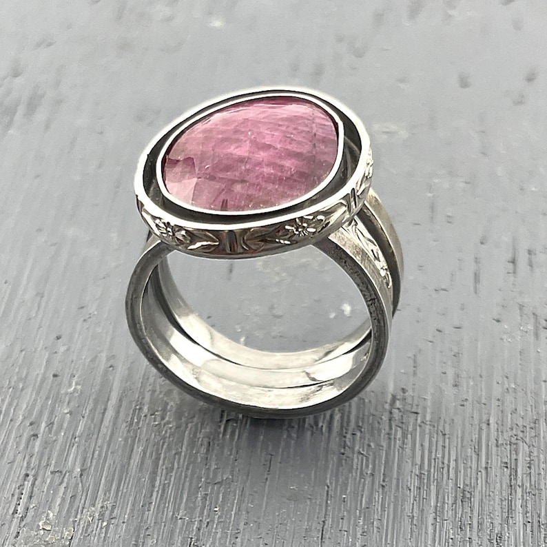 May include: A silver-toned ring featuring a large, faceted, oval-shaped pink gemstone set within a decorative bezel. The band consists of two distinct silver-colored rings, with the upper portion displaying an engraved floral pattern around the stone.