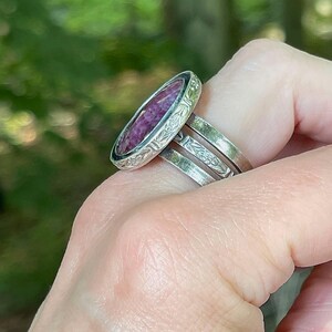 May include: A silver ring featuring an oval-shaped, deep magenta-colored stone set within a decorative bezel. The band consists of three distinct silver segments, with the middle segment displaying an engraved floral pattern along its outer surface.
