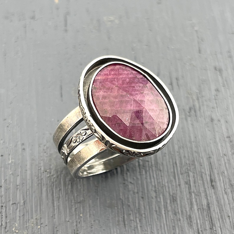 May include: A silver ring featuring a large, oval, faceted magenta-pink gemstone set in a dark metal bezel. The band consists of three stacked silver layers, with the outer bands displaying a delicate, embossed floral pattern along the edges.