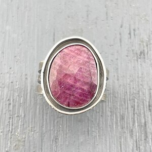 May include: A ring featuring a large, oval-shaped, faceted pinkish-red gemstone set within a smooth, polished silver-toned metal bezel. The band of the ring is wide and constructed from multiple layered silver-toned metal strips with a textured finish.