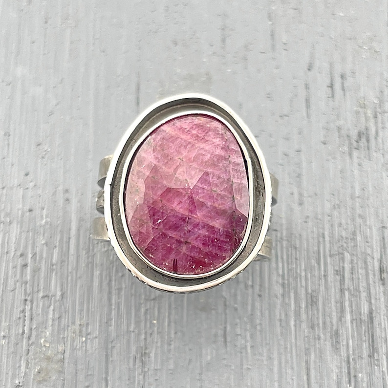 May include: A ring featuring a large, oval-shaped, faceted pinkish-red gemstone set within a smooth, polished silver-toned metal bezel. The band of the ring is wide and constructed from multiple layered silver-toned metal strips with a textured finish.