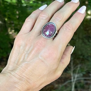 May include: A silver ring featuring a large, oval-shaped, faceted magenta gemstone set in a smooth, polished metal bezel. The band of the ring is constructed from multiple thin, parallel silver wires that connect to the central stone setting.