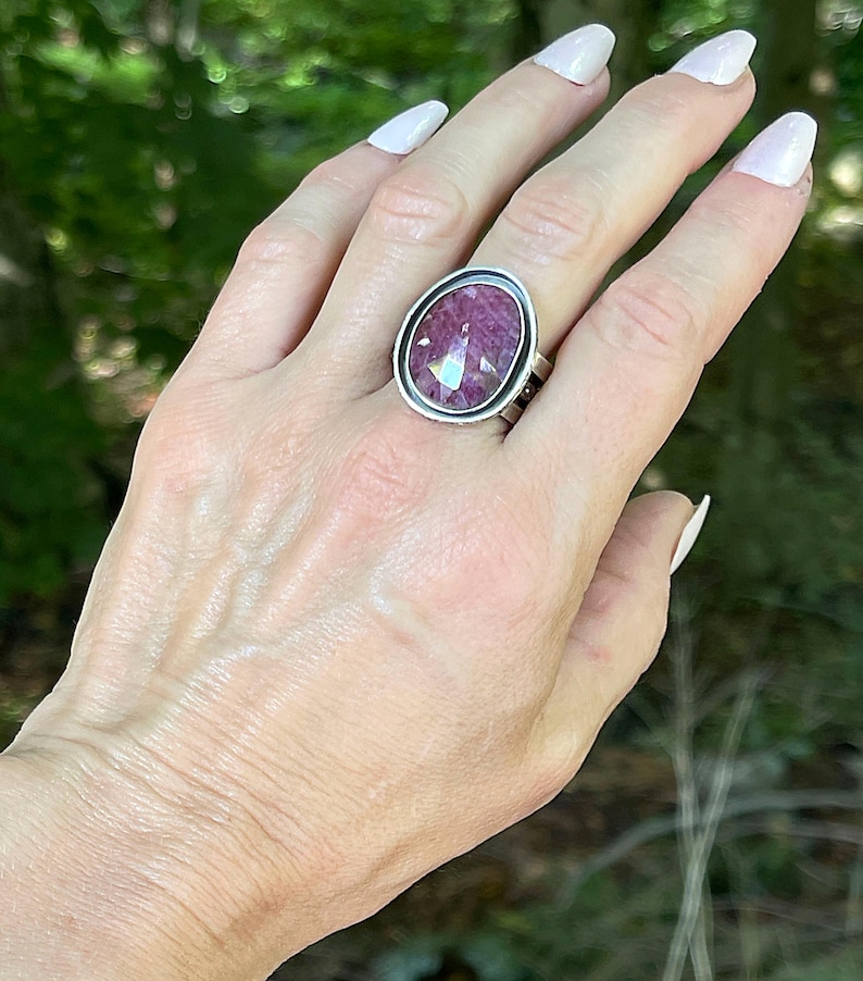 May include: A silver ring featuring a large, oval-shaped, faceted magenta gemstone set in a smooth, polished metal bezel. The band of the ring is constructed from multiple thin, parallel silver wires that connect to the central stone setting.