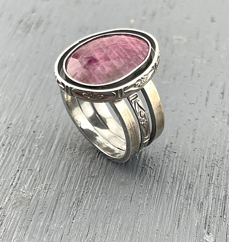May include: A silver ring featuring a large, oval-shaped, faceted pink gemstone set in a bezel. The ring band consists of three distinct silver components, with the top section displaying an engraved floral pattern and a darkened, oxidized finish.