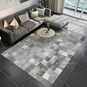 Puede incluir: Una alfombra con estampado geométrico gris y blanco con el texto "Eclatigue Rug" se muestra en un salón. La alfombra se coloca frente a un sofá seccional gris con cojines decorativos y una mesa de centro redonda de madera.