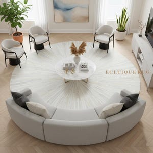 Może przedstawiać: Nowoczesny układ salonu z jasnoszarą sofą modułową, czterema fotelami i okrągłym stolikiem kawowym. Centralnym punktem jest duży, okrągły dywan z wzorem promieni słonecznych. Pokój jest udekorowany roślinami i obrazem.