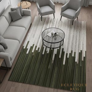 Op de afbeelding: Een modern vloerkleed met een geometrisch ontwerp. Het vloerkleed heeft een gradiëntpatroon, dat overgaat van donkergroen aan de onderkant naar witte en grijze strepen. De tekst "ECLECTIQUE RUG" is zichtbaar op het vloerkleed.