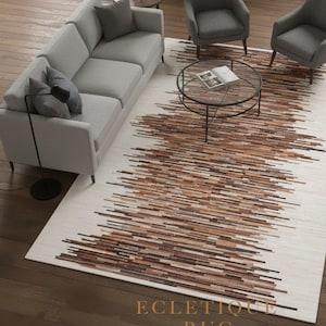 Peut inclure: Un tapis rectangulaire crème avec un motif de rayures horizontales marron et beige. Le tapis est dans un salon avec un canapé gris, des chaises et une table basse en verre. Le texte "ECLECTIQUE RUG" est visible.