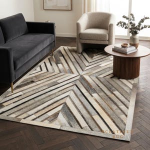 Peut inclure: Un tapis rectangulaire composé de bandes de cuir cousues dans des tons crème, beige, gris et brun foncé. Le motif présente des lignes diagonales se rejoignant en points centraux, créant un effet de chevron multidirectionnel sur toute la surface.