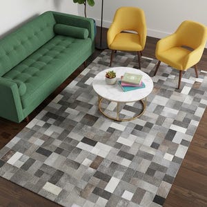 Op de afbeelding: Een woonkamer met een groene bank, twee gele stoelen en een ronde salontafel. Een geometrisch patroon tapijt in grijstinten, wit en bruin bedekt de vloer. Een vloerlamp en een potplant zijn ook zichtbaar.