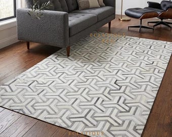 May include: A rectangular area rug featuring a repeating geometric pattern of interlocking Y-shaped forms in shades of white, light grey, and dark grey. The rug is positioned on a dark wood floor, partially underneath a grey sofa and a lounge chair.