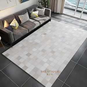 Puede incluir: Una alfombra rectangular grande con un patrón geométrico de cuadrados grises claros y blancos. La alfombra está colocada sobre un suelo de baldosas gris oscuro, con un sofá gris y cojines decorativos en el fondo. El texto "ECLATIQUE RUG" es visible.