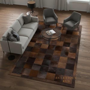 Puede incluir: Una alfombra rectangular de patchwork marrón y negro, con un borde tachonado. La alfombra, con el texto "ECLECTIQUE RUG", está colocada en un salón y presenta un patrón geométrico de cuadrados. La habitación incluye un sofá y sillones.