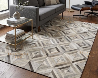 May include: A rectangular area rug featuring a geometric pattern of interlocking squares and triangles in shades of grey, white, and beige. The rug is composed of stitched panels with a textured surface, displayed in a room with a sofa and chair.