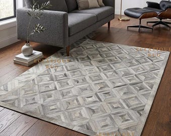 May include: A rectangular area rug featuring a geometric pattern of stitched grey and white hide patches. The rug has a repeating square design with a solid border. The text ECLATIQUE RUG is visible in gold lettering across the surface of the rug.
