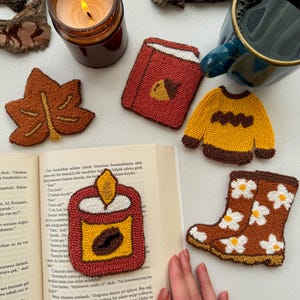 May include: An assortment of handmade, embroidered items, including a leaf, a book, a sweater, a candle, and a boot with floral accents. A lit candle and a mug of coffee are also present, suggesting a cozy, autumn theme.