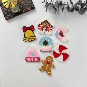 May include: A collection of colorful, embroidered Christmas-themed patches. Designs include a yellow bell with a red bow, a gingerbread house, a pink stocking, a snow globe with a green tree, a pink hat, a gingerbread man, and a red and white peppermint candy.
