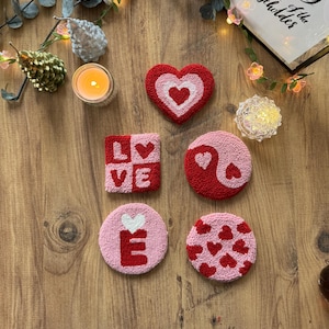 May include: A collection of handmade, textured coasters in various shapes and designs. The coasters feature heart, yin-yang, and circular shapes, with the word "LOVE" and the letter "E" incorporated. The color palette includes red, pink, and white, set against a wooden surface.