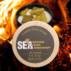 May include: A round wooden lid with the text "Campfire S'more Escape" and "SEA SURVIVORS ESCAPE AROMATHERAPY" sits in front of a black container with green and white candle wax. The label reads "All Natural 4 oz./113 g Made in VA".