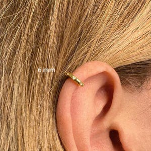 May include: A gold-colored ear cuff with three small, clear gemstones. The cuff is 6 mm in size and is worn on the ear. The background shows blonde hair.
