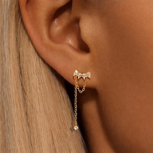 May include: Gold earring featuring a bow-shaped top set with three clear, teardrop-shaped stones. A fine chain hangs from the bow, ending in a single, sparkling stone. The earring is designed to be worn on the ear.